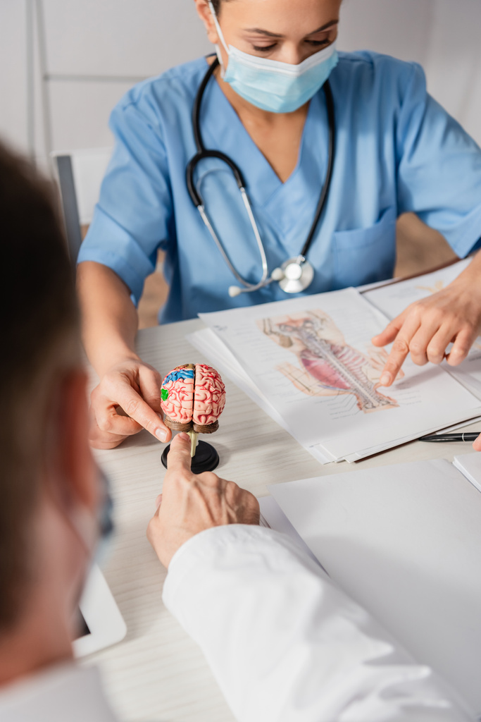 Wieloetniczny personel szpitalny wskazujący palcami na anatomiczny model mózgu i obraz w miejscu pracy na niewyraźnym pierwszym planie - Zdjęcie, obraz
