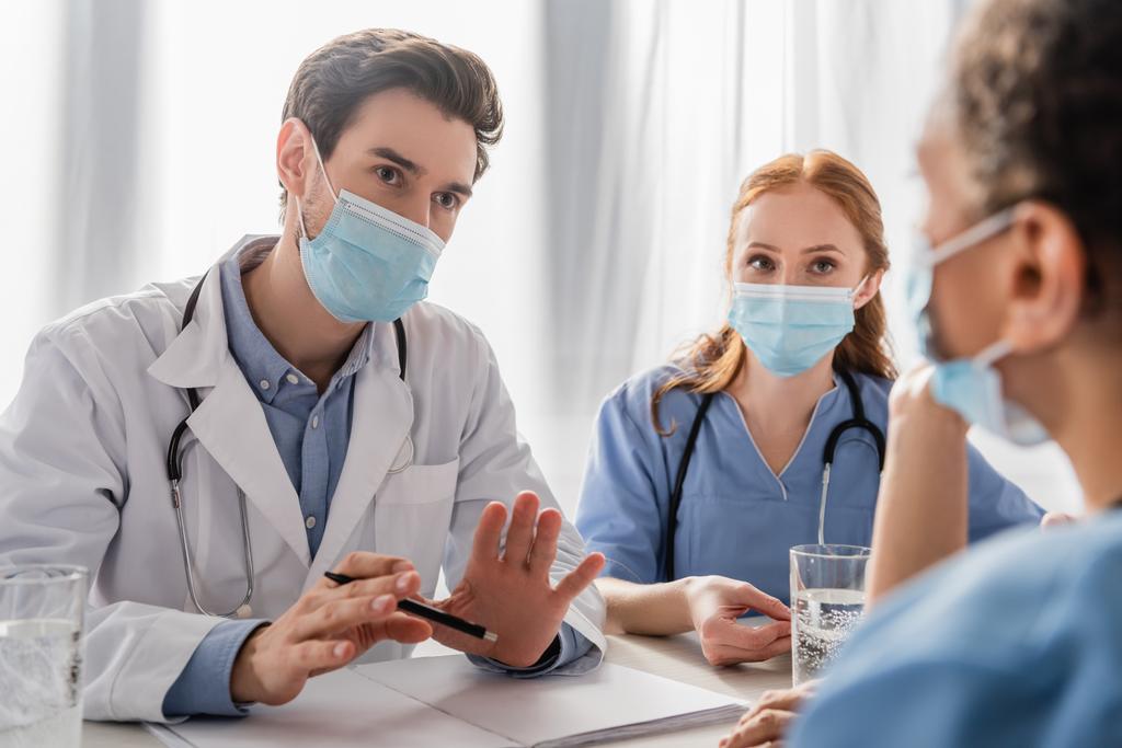 Врач в медицинской маске сидит рядом с медсестрой и разговаривает с африканским американским коллегой на рабочем месте на размытом переднем плане - Фото, изображение