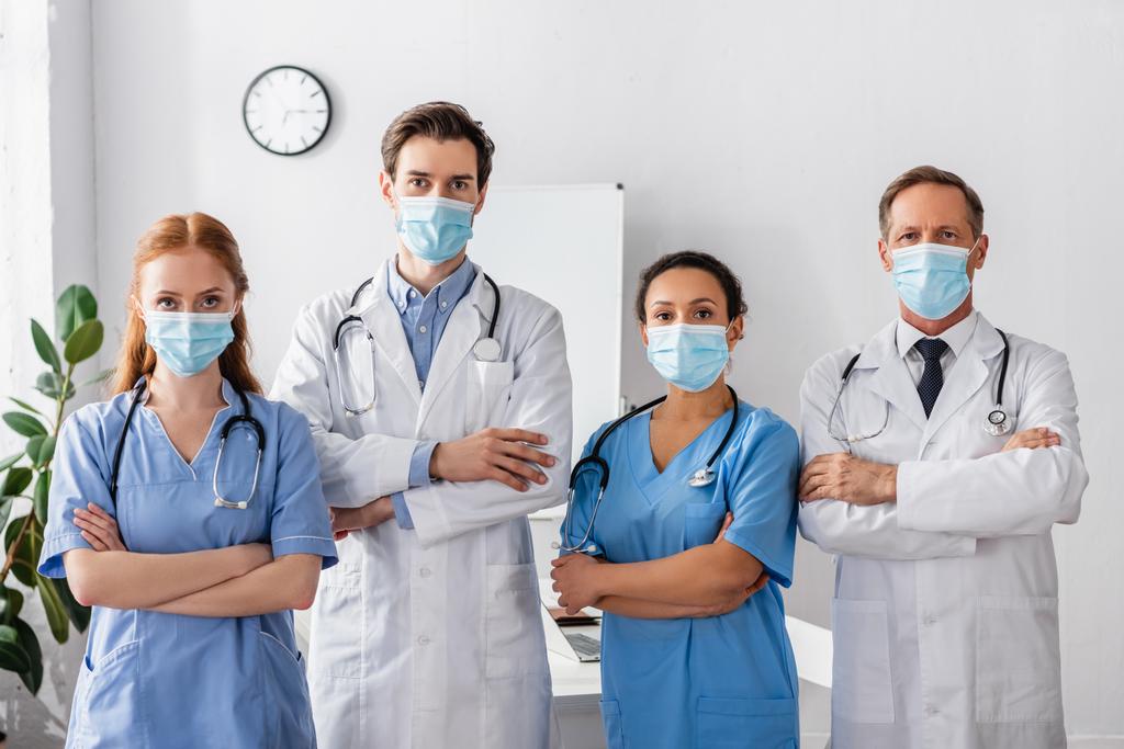 Tıp maskeli çok kültürlü doktorlar ve hemşireler hastanede yan yana dururken kameraya bakıyorlar. - Fotoğraf, Görsel