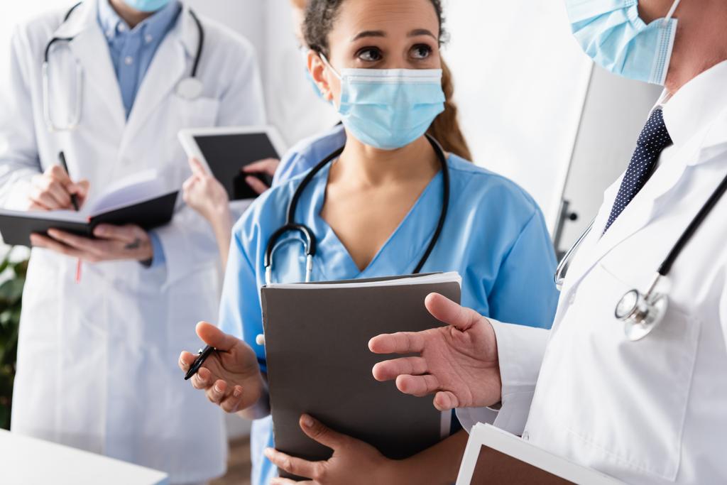 Afrikanisch-amerikanische Krankenschwester in medizinischer Maske im Gespräch mit Arzt mit verschwommenen Kollegen im Hintergrund - Foto, Bild