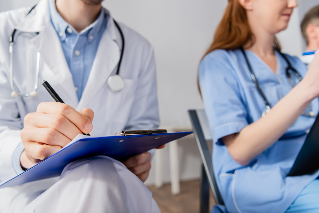 Przycięty widok lekarza z stetoskopem na schowku, siedzącego obok pielęgniarki podczas spotkania na niewyraźnym tle - Zdjęcie, obraz