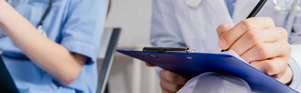 Καλλιεργημένη άποψη του γιατρού γράφοντας στο πρόχειρο κοντά στο συνάδελφο κατά τη διάρκεια συνεδρίασης στο νοσοκομείο σε θολή φόντο, πανό - Φωτογραφία, εικόνα