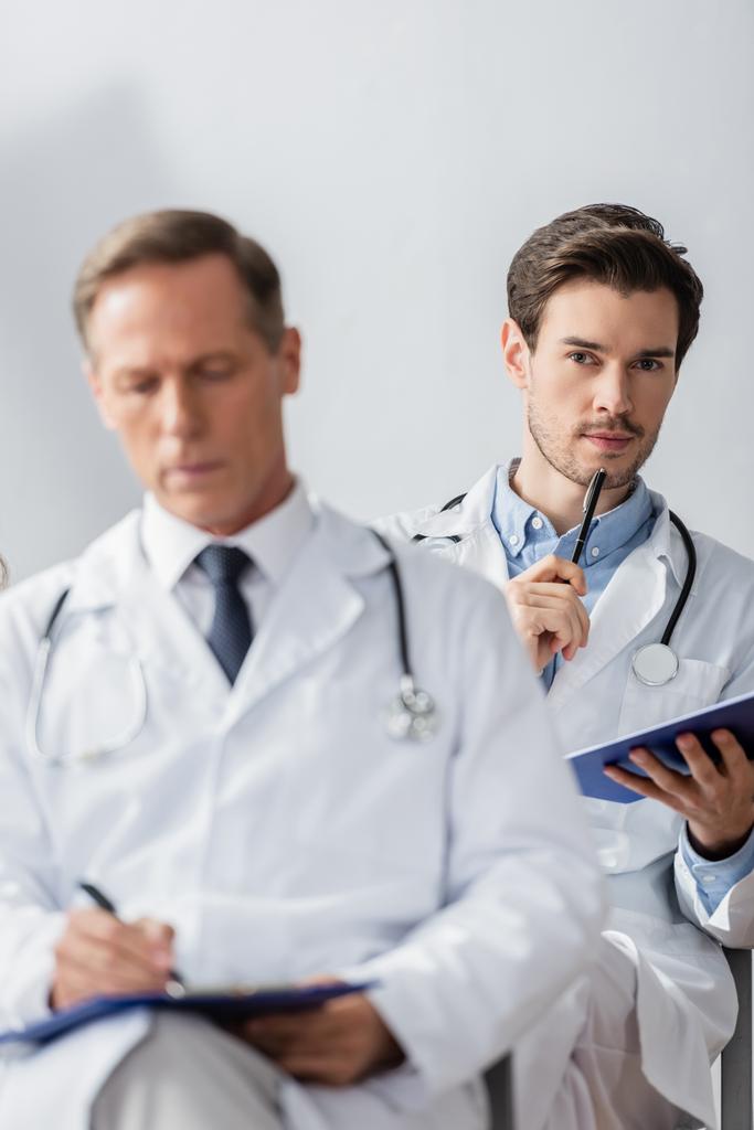 Bedachtzame arts met pen en klembord kijkt naar camera tijdens ontmoeting met wazige collega op de voorgrond in het ziekenhuis - Foto, afbeelding