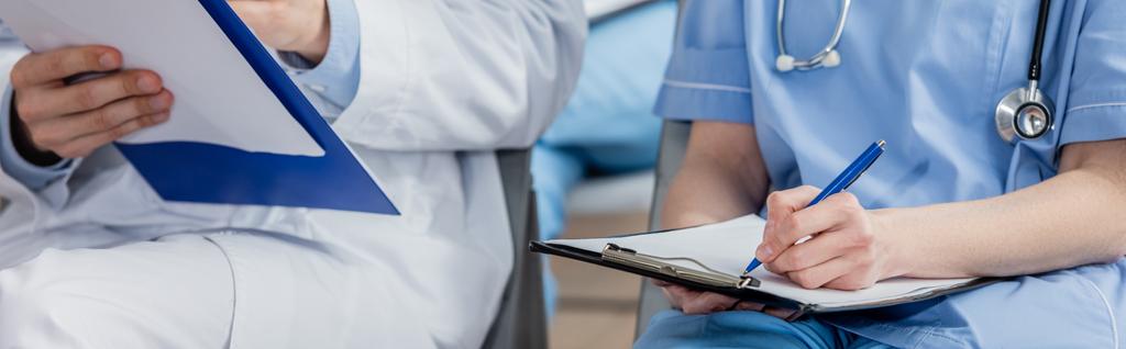 Nurse writing on clipboard while sitting near doctor during meeting in hospital on blurred background, banner - Photo, Image