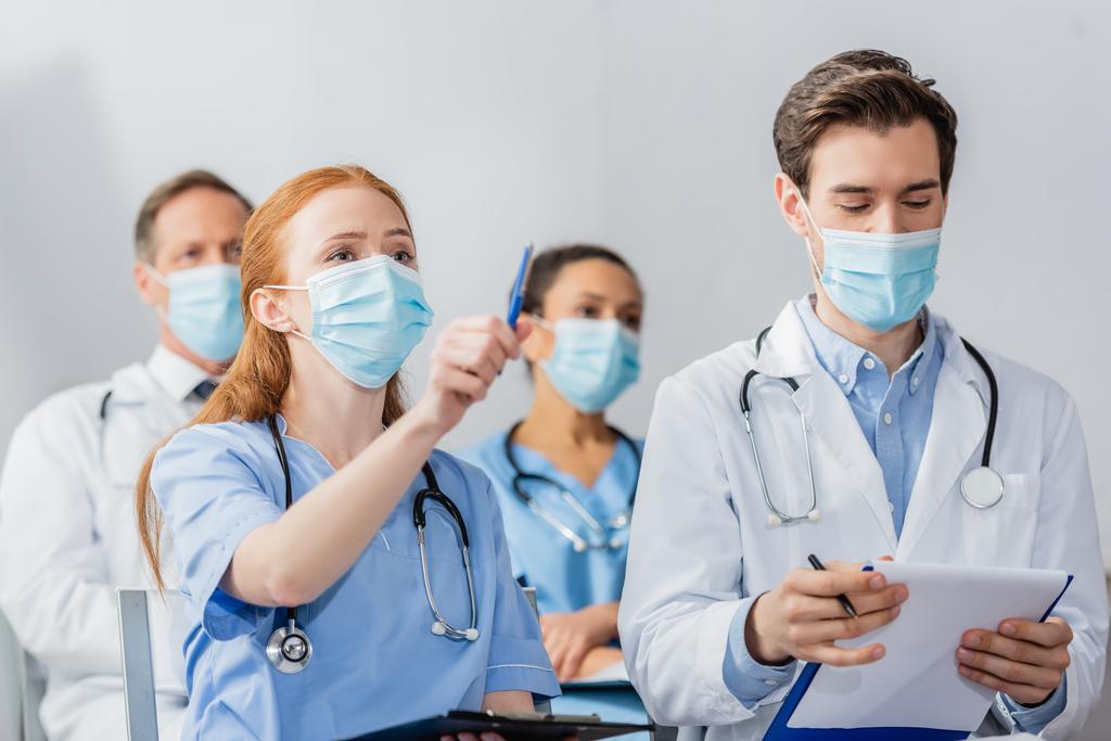 Рыжая медсестра указывает ручкой, сидя рядом с врачом во время встречи с размытыми коллегами на заднем плане в больнице - Фото, изображение