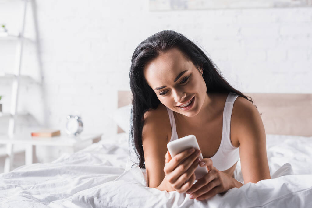 Lächelnde junge brünette Frau im Bett mit Smartphone - Foto, Bild