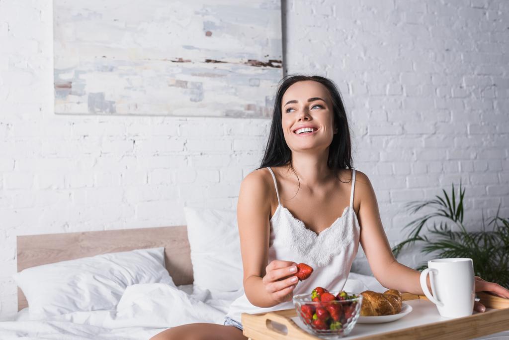 glückliche junge brünette Frau mit Croissant und Erdbeere zum Frühstück im Bett - Foto, Bild