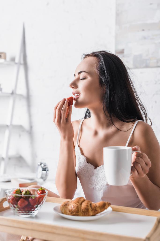 若いブルネットの女性がベッドで朝食のためにイチゴを食べる - 写真・画像
