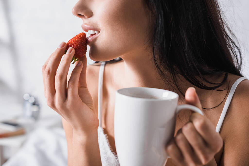 abgeschnittene Ansicht einer jungen brünetten Frau mit Becher, die Erdbeere zum Frühstück isst - Foto, Bild