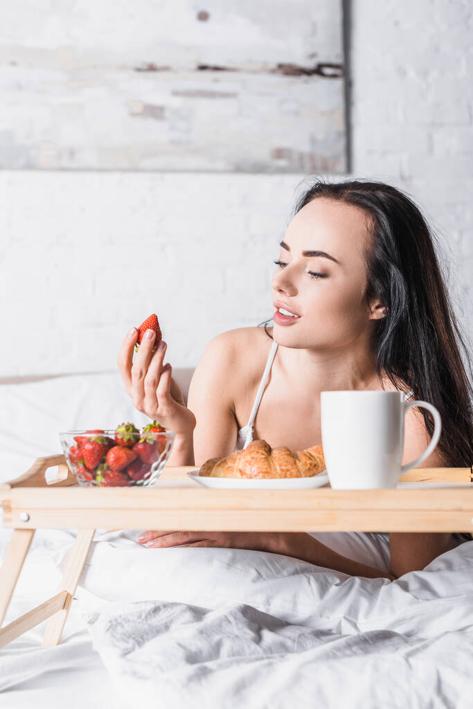 Genç esmer kadın kahvaltıda kruvasan ve çilek yiyor ve yatakta kakao içiyor. - Fotoğraf, Görsel