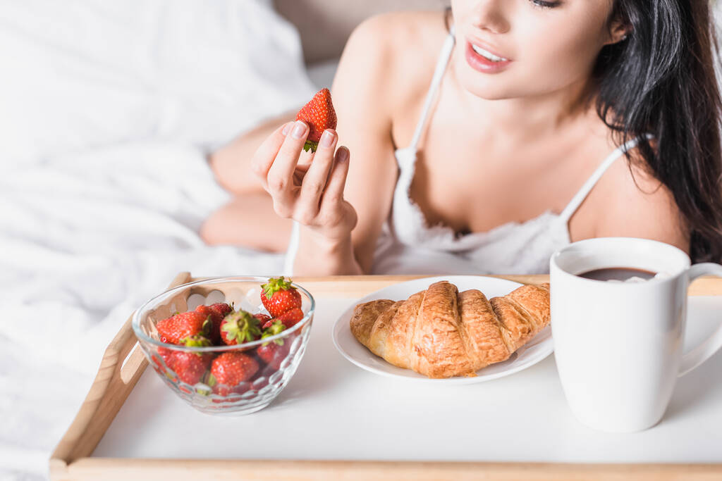 vista cortada de jovem morena que tem croissant e morango para o café da manhã e beber cacau na cama - Foto, Imagem