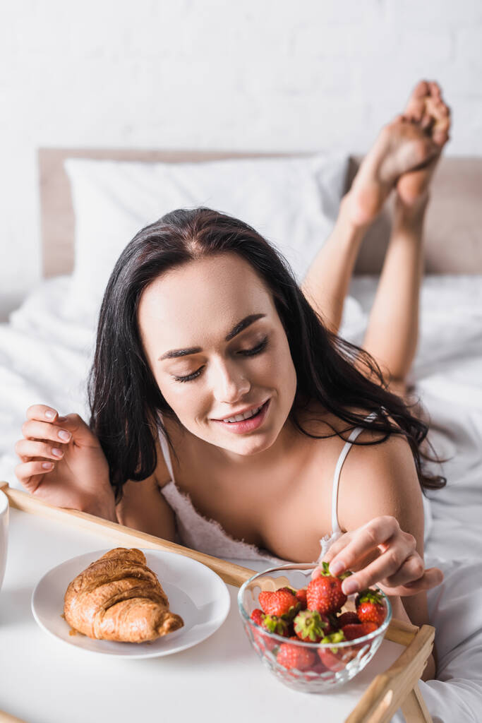 nuori brunette nainen ottaa croissant, mansikka aamiaiseksi sängyssä - Valokuva, kuva