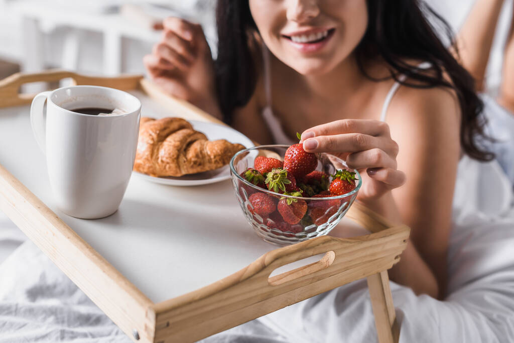 vista recortada de la joven morena tomando croissant, fresa y cacao para el desayuno en la cama - Foto, Imagen