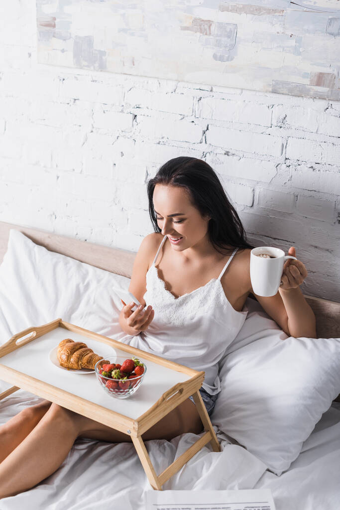 νεαρή μελαχρινή γυναίκα έχει κρουασάν, φράουλα και κακάο για πρωινό, ενώ χρησιμοποιώντας smartphone στο κρεβάτι - Φωτογραφία, εικόνα
