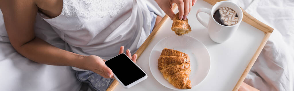 vista recortada de la mujer que tiene croissant y cacao para el desayuno mientras usa el teléfono inteligente en la cama, pancarta - Foto, imagen
