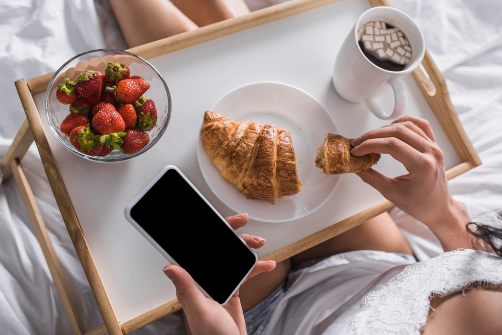 oříznutý pohled na ženu s croissantem, jahodami a kakaem k snídani při držení smartphonu v posteli - Fotografie, Obrázek