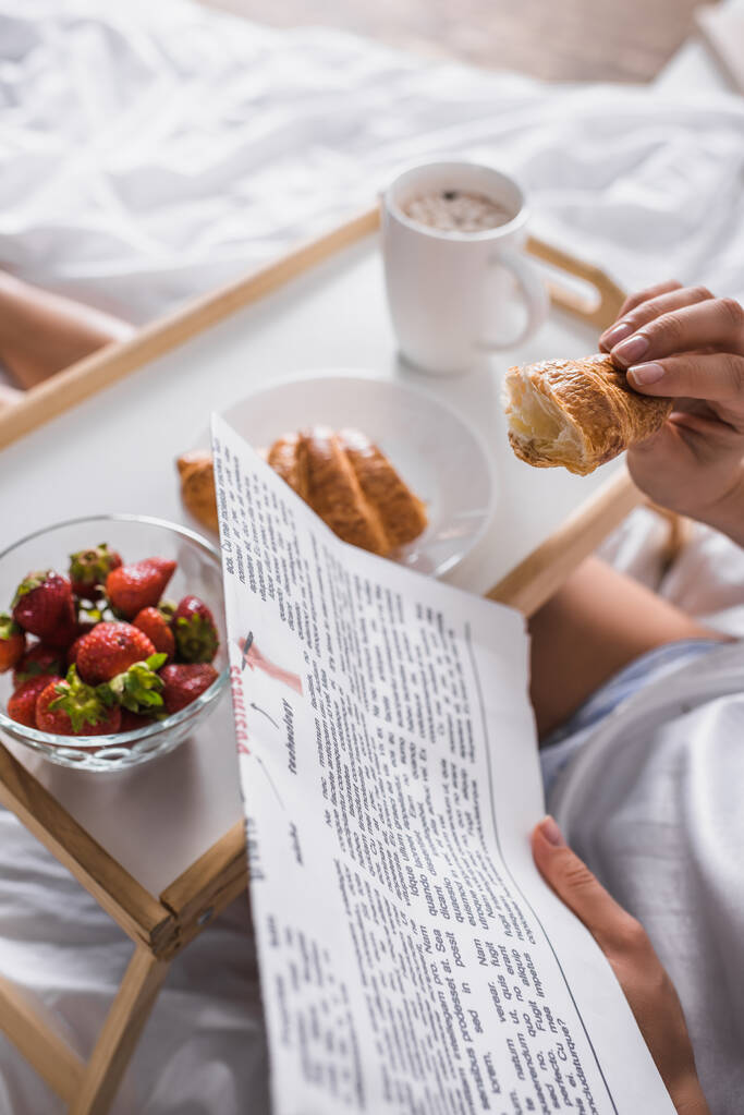 καλλιεργημένη άποψη της γυναίκας που έχει κρουασάν, φράουλα και κακάο για πρωινό, ενώ διαβάζετε εφημερίδα στο κρεβάτι - Φωτογραφία, εικόνα