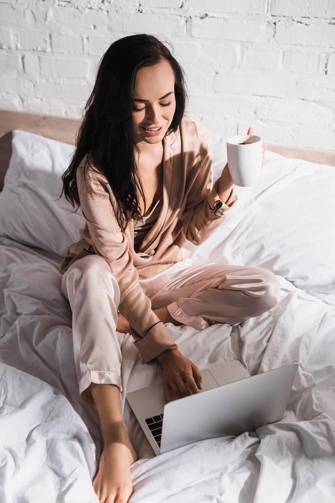 νεαρή μελαχρινή γυναίκα κάθεται στο κρεβάτι με κούπα και φορητό υπολογιστή το πρωί - Φωτογραφία, εικόνα