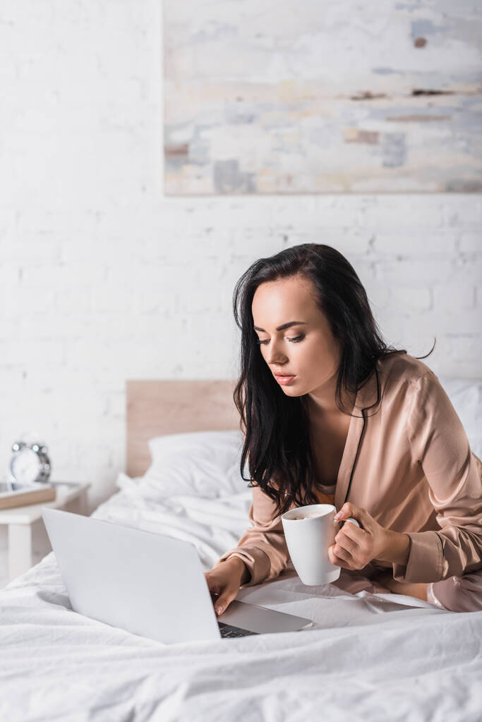 junge brünette Frau sitzt morgens mit Becher und Laptop im Bett - Foto, Bild