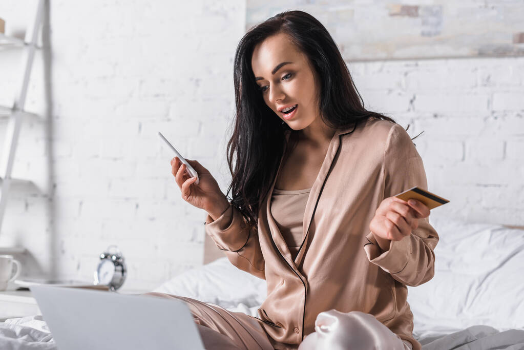 zszokowana młoda brunetka siedzi rano w łóżku ze smartfonem, kartą kredytową i laptopem - Zdjęcie, obraz
