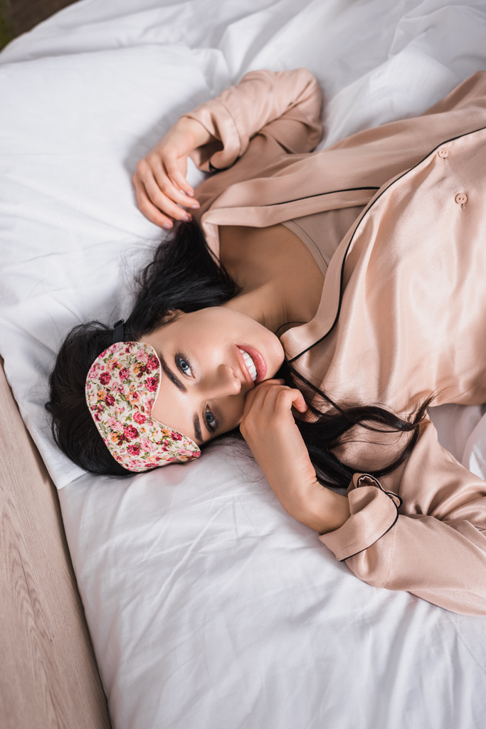 vista superior de la joven morena sonriente tumbada en la cama en la máscara del sueño - Foto, imagen