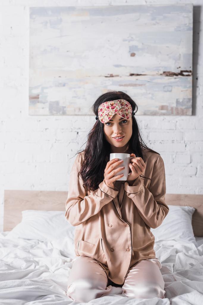 young brunette woman sitting in bed in sleep mask with mug of cocoa - Photo, Image