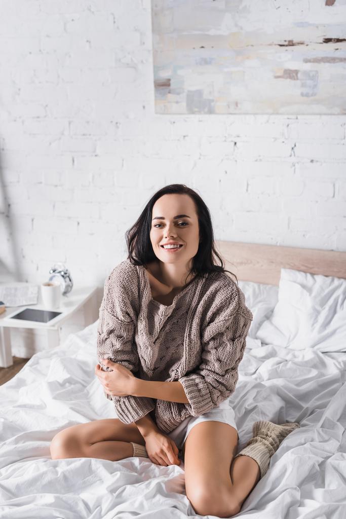 Ευτυχισμένη νεαρή μελαχρινή γυναίκα με πουλόβερ και κάλτσες που κάθεται στο κρεβάτι το πρωί - Φωτογραφία, εικόνα