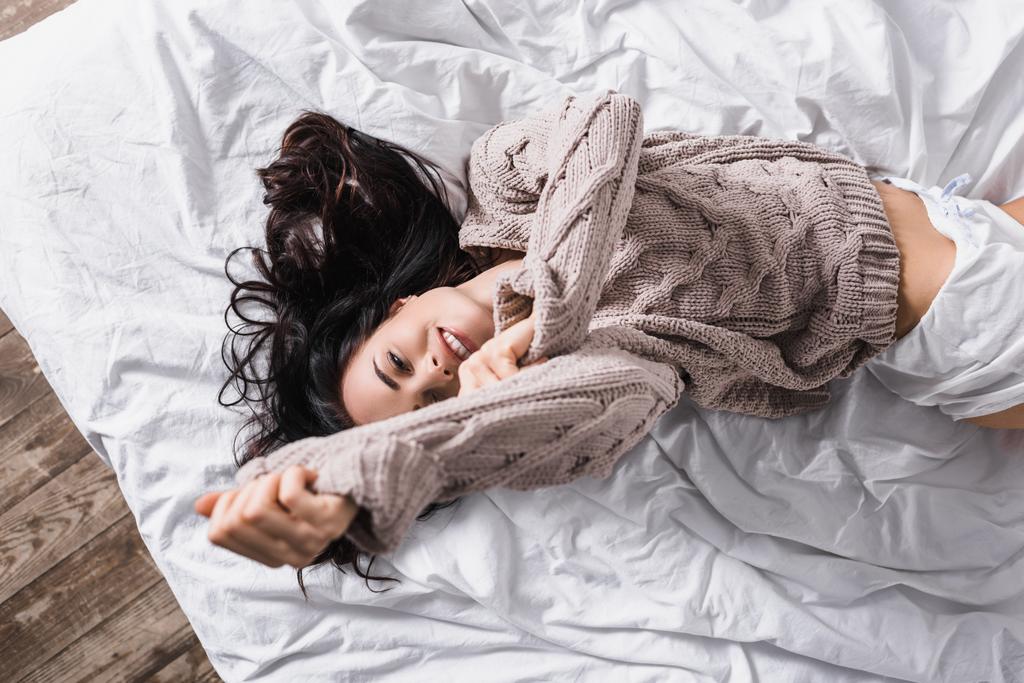 top uitzicht van glimlachende jonge brunette vrouw in trui liggend in bed in de ochtend - Foto, afbeelding