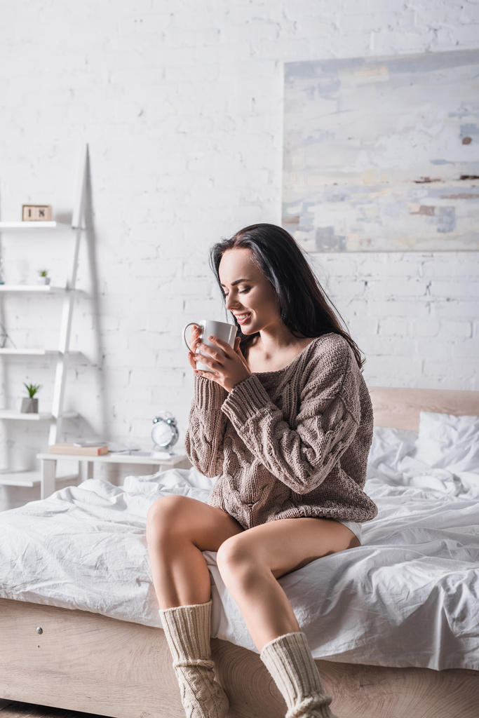 νεαρή μελαχρινή γυναίκα σε πουλόβερ κάθεται στο κρεβάτι με κούπα ζεστό κακάο το πρωί - Φωτογραφία, εικόνα