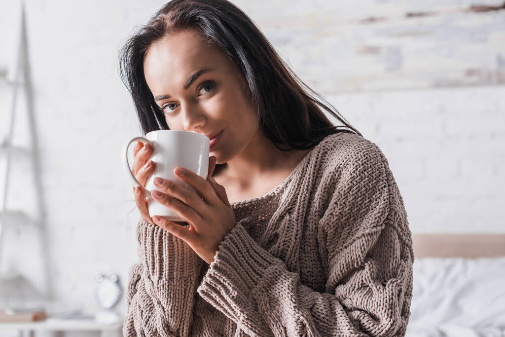 giovane donna bruna in maglione seduta a letto con tazza di cioccolata calda al mattino - Foto, immagini