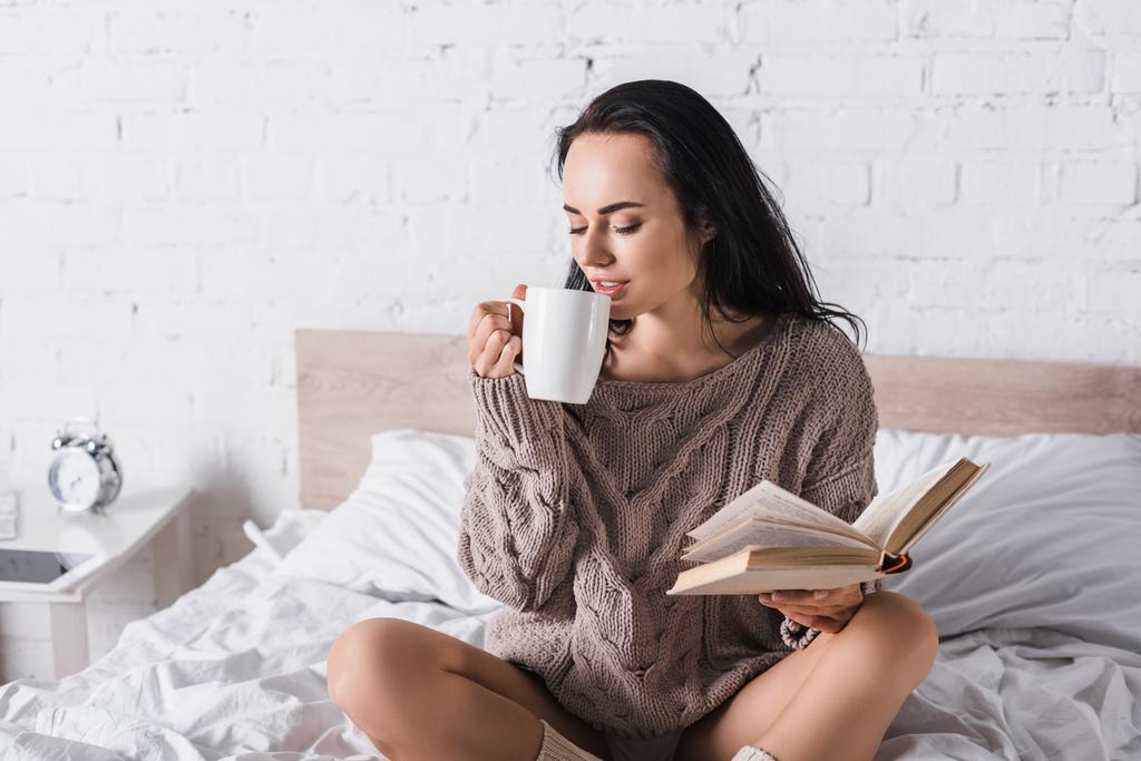 junge brünette Frau im Pullover sitzt im Bett mit einem Becher heißen Kakaos und Buch am Morgen - Foto, Bild