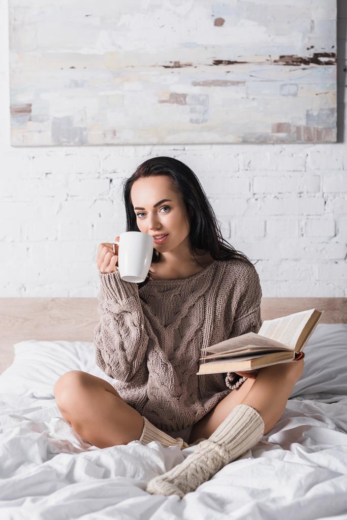 jonge brunette vrouw in trui zitten in bed met mok van warme chocolademelk en boek in de ochtend - Foto, afbeelding
