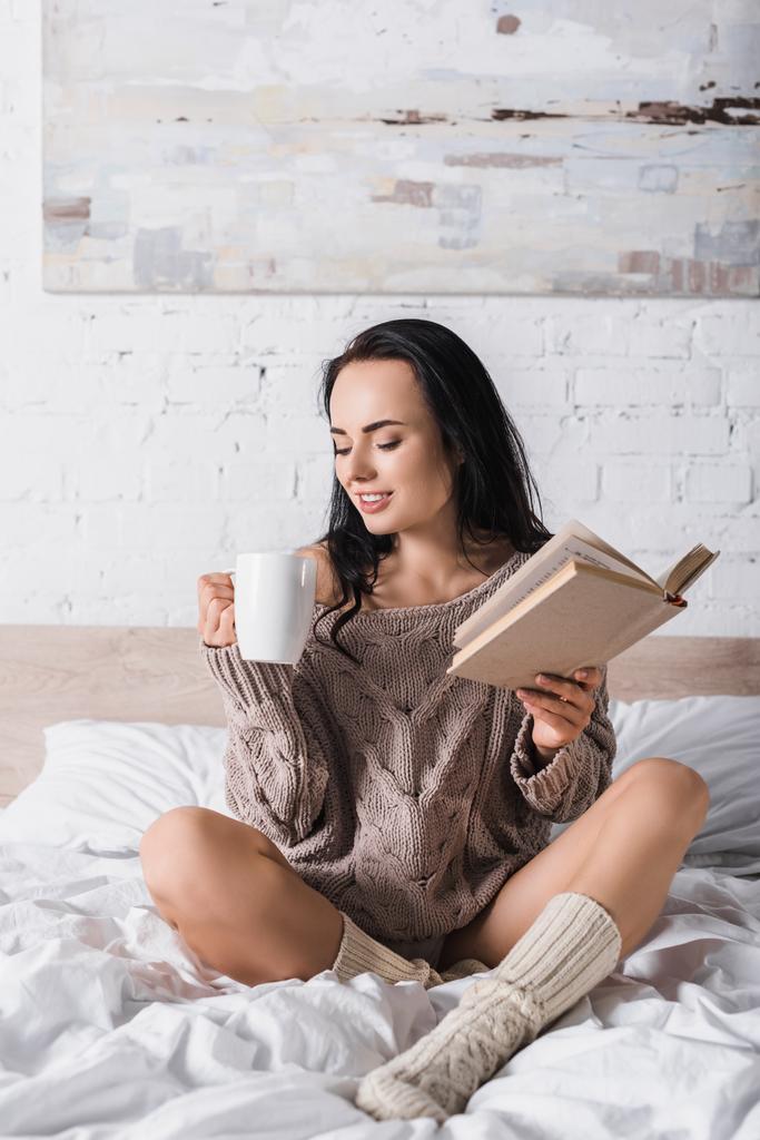 glimlachende jonge brunette vrouw in trui zitten in bed met mok warme chocolademelk en boek in de ochtend - Foto, afbeelding