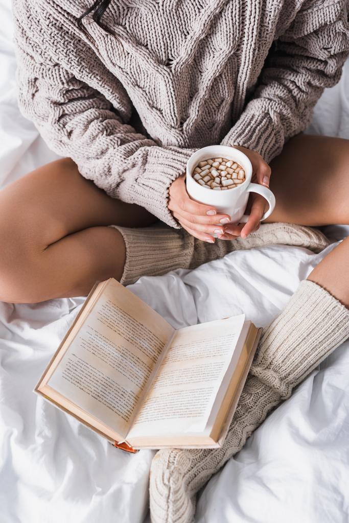 vue recadrée de la femme en pull et chaussettes tricotées assis dans le lit avec tasse de cacao chaud et livre le matin - Photo, image