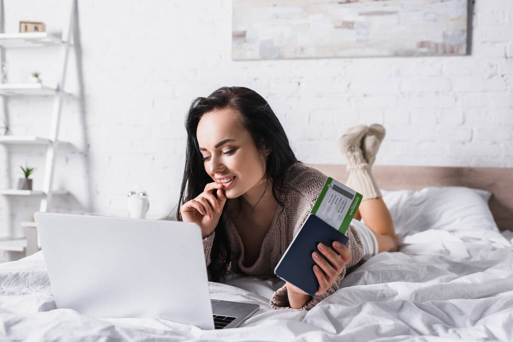 sorridente giovane donna bruna in maglione sdraiata a letto con laptop e biglietto aereo - Foto, immagini