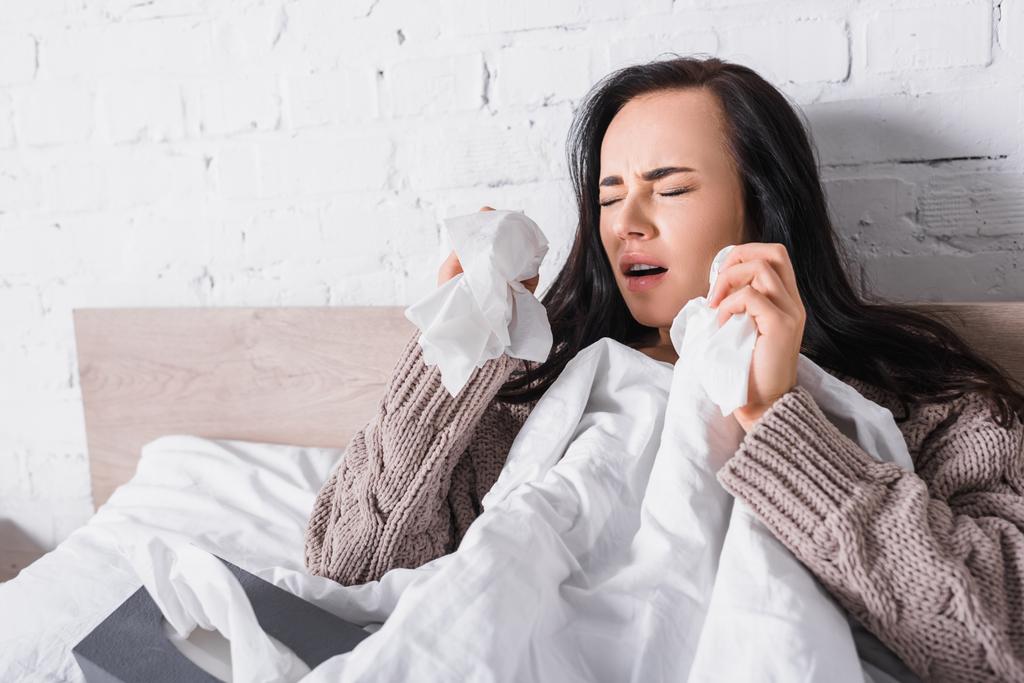 enferma joven morena mujer en suéter estornudando en la cama con tejido - Foto, Imagen