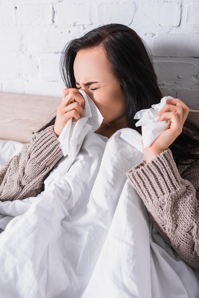 enferma joven morena mujer en suéter estornudando en la cama con tejido - Foto, imagen