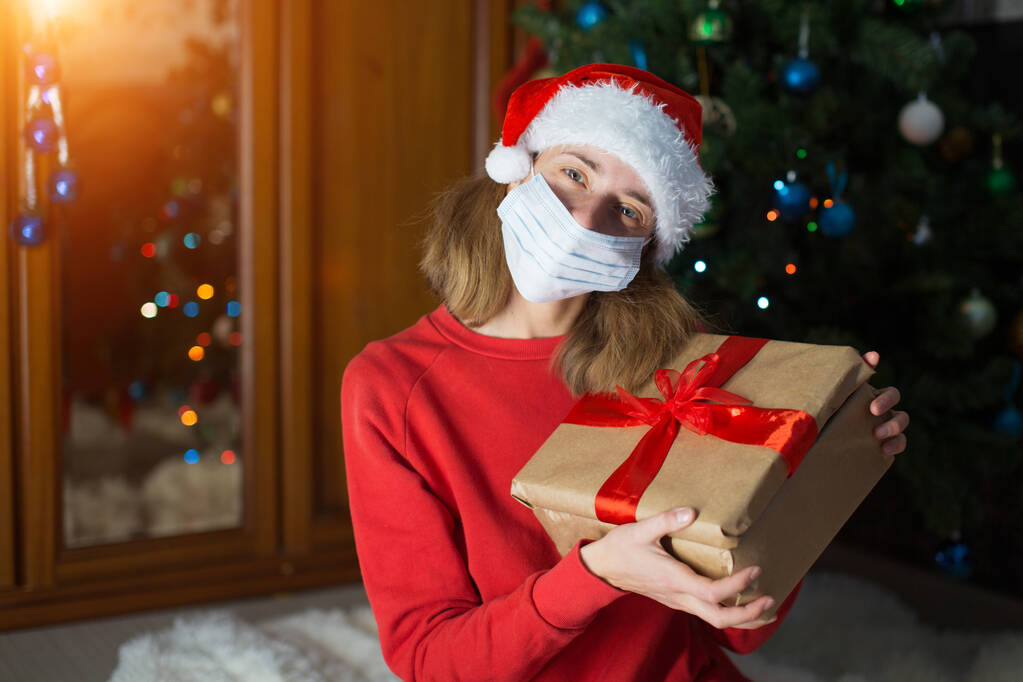 κορίτσι με κόκκινα ρούχα και ιατρική μάσκα ξεδιπλώνουν τα δώρα της Πρωτοχρονιάς. την ιδέα του εορτασμού των Χριστουγέννων τα μεσάνυχτα. Διακόσμηση των διακοπών boce, Χριστουγεννιάτικο δέντρο - Φωτογραφία, εικόνα