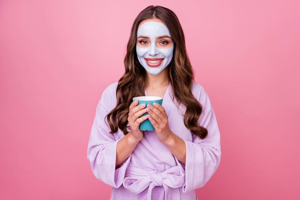 Foto retrato de menina bonita fazendo procedimentos de tratamento para a pele usando máscara facial segurando xícara de café vestindo roupão isolado no fundo cor-de-rosa - Foto, Imagem