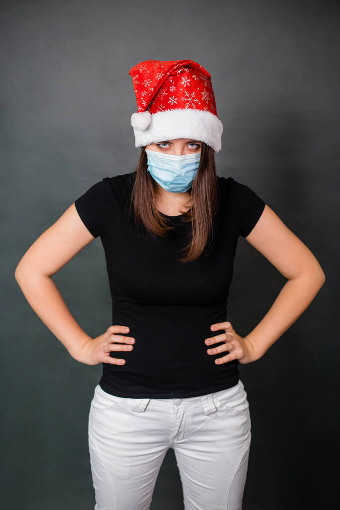 Egy fekete pólós nő portréja, egy eldobható orvosi maszk és egy szürke hátterű karácsonyi kalap. Egyedül karantén karácsonyi ünneplés koncepció. - Fotó, kép