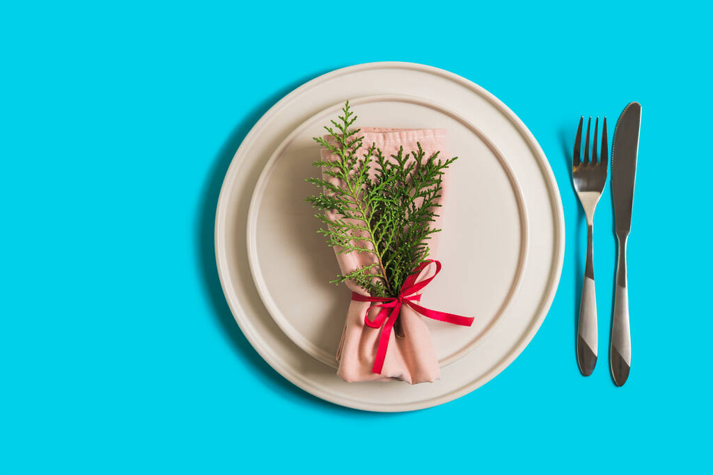 Geserveerd tafel met leeg bord en bestek voor de viering van Kerstmis en Nieuwjaar. Op het bord ligt servet met kerstboomtak. Flatlay op helderblauwe achtergrond. Bovenaanzicht. - Foto, afbeelding