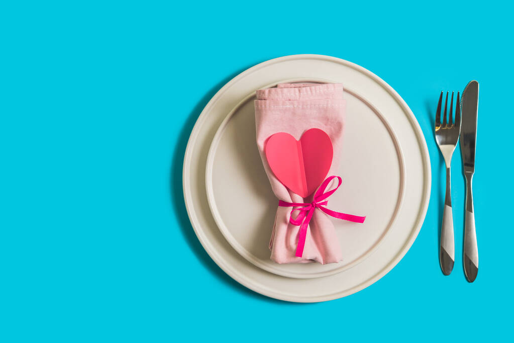 Served table with plate and cutlery for the celebration of Valentines Day. On the plate is napkin with paper heart. Flatlay on bright blue background. Top view. - Photo, Image