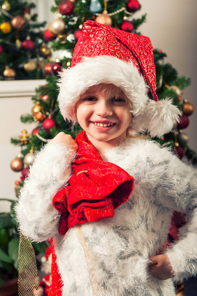 Bella bambina con cappello di Babbo Natale e sacchetto regalo accanto all'albero di Natale - Foto, immagini