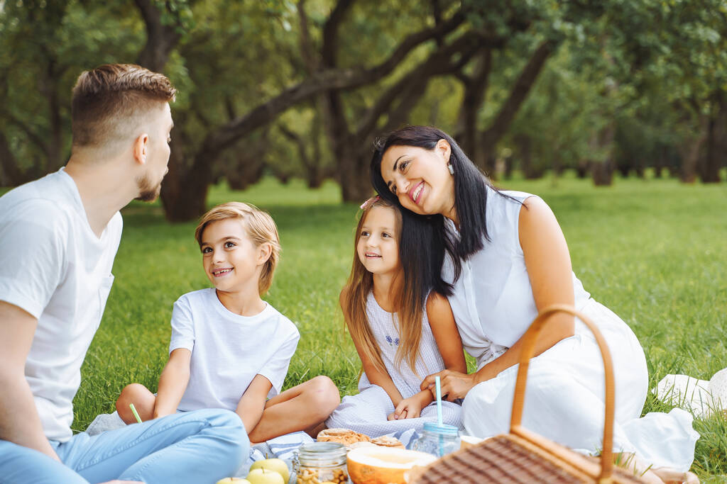 Szczęśliwa rodzina z dwójką dzieci relaksujących się na trawniku podczas pikniku w zielonym ogrodzie. - Zdjęcie, obraz