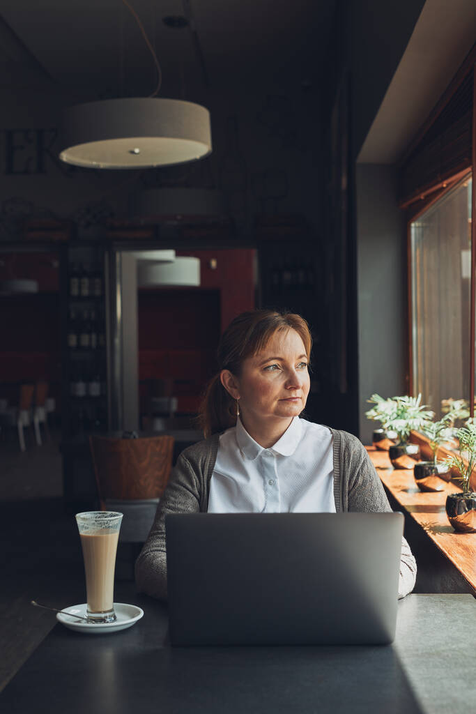Entspannte Geschäftsfrau, die ferngesteuert an ihrem Laptop arbeitet, erwägt, ihre Arbeit am Tisch in einem Café zu erledigen - Foto, Bild