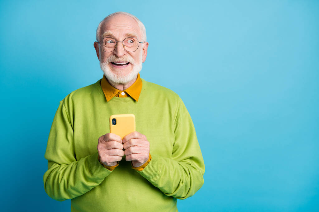 Foto del abuelo retirado mantenga el lado del aspecto del teléfono Espacios vacíos use especificaciones suéter verde aislado fondo de color azul - Foto, imagen