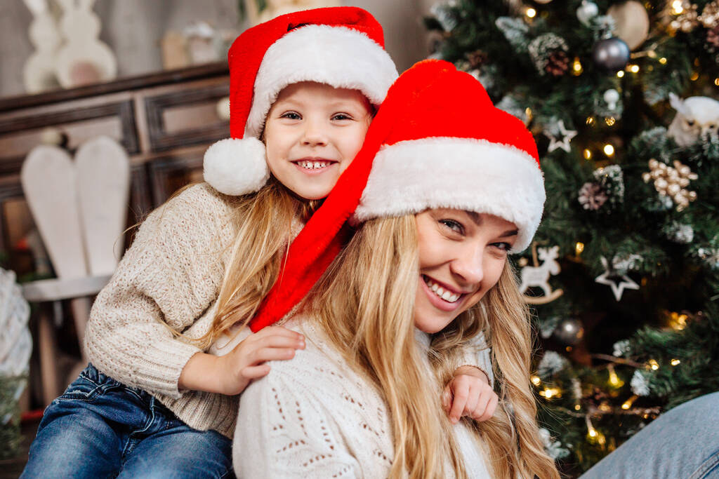 Portrait of happy mother in santa claus and daughter near christmas tree at home. Merry Christmas - Photo, Image