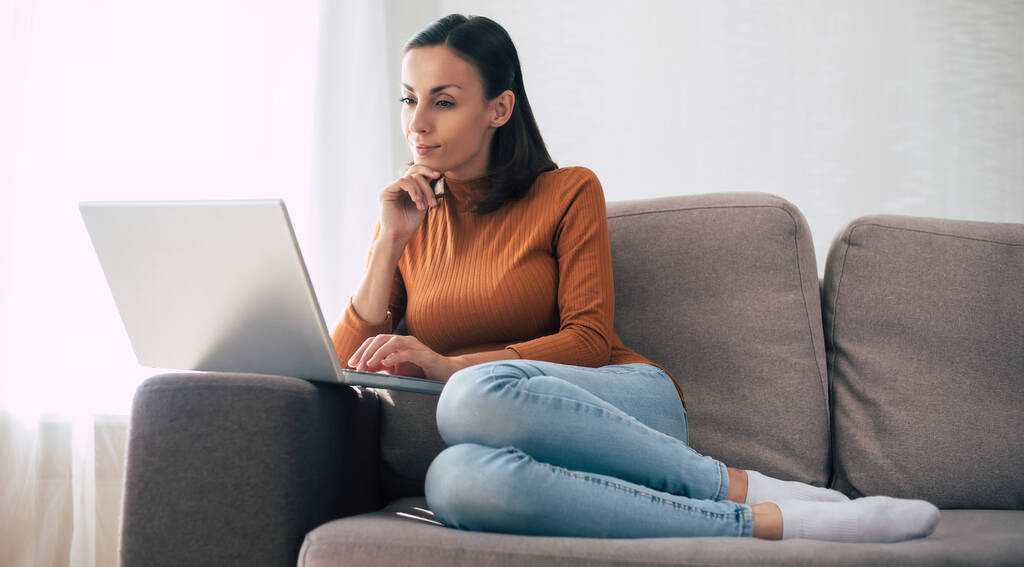 Молода впевнена красива жінка працює зі своїм ноутбуком, сидячи на дивані вдома
 - Фото, зображення