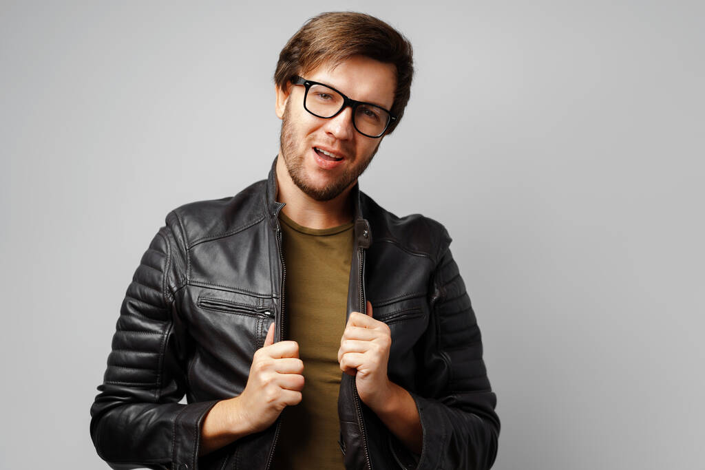 Portrét mladého muže v brýlích v černé kožené bundě na šedém pozadí - Fotografie, Obrázek