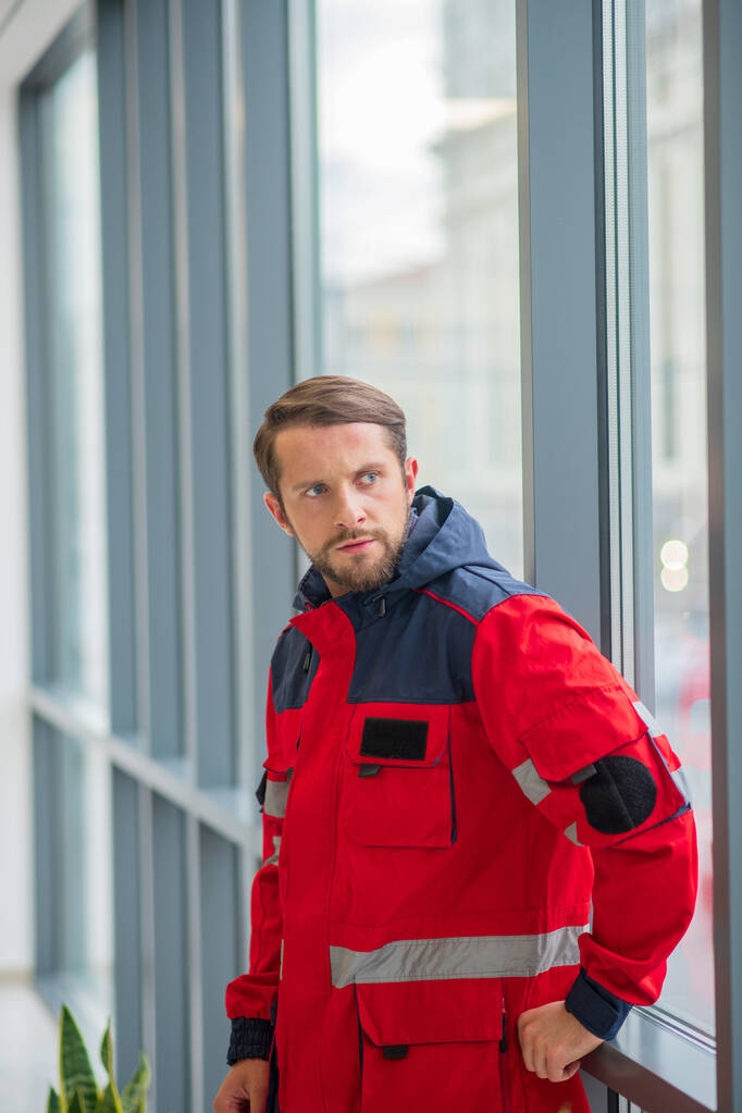 Muž doktor v červené uniformě stojí u okna a vypadá ustaraně - Fotografie, Obrázek